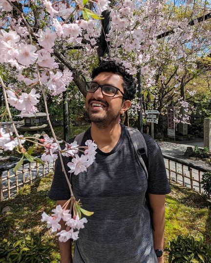 A photo of Mahesh Makani, under a cherry blossom tree. He is smiling and looking towards the top left of the image.