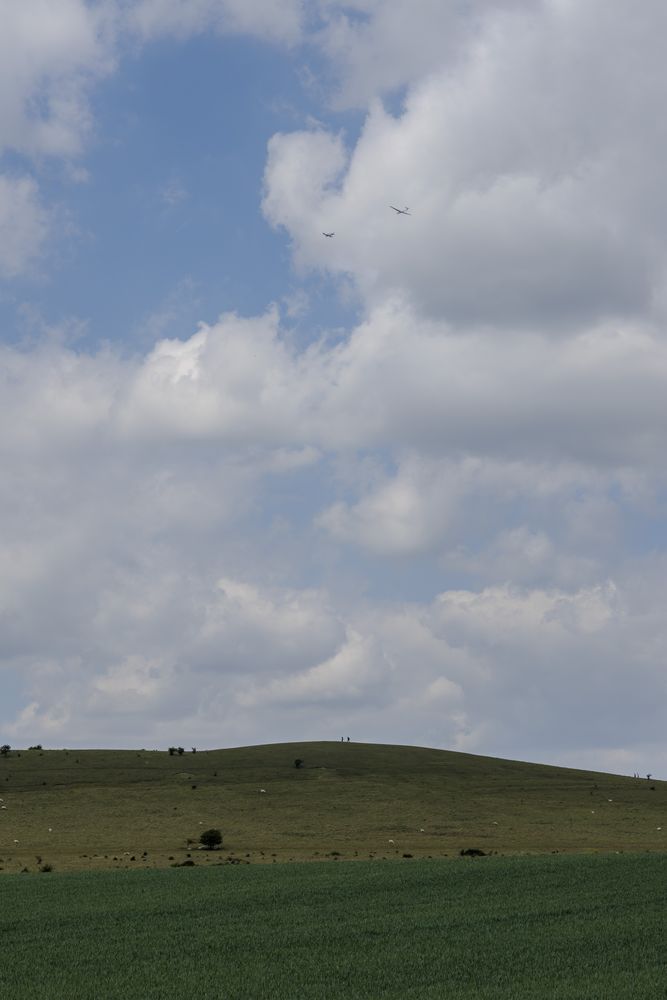 A glider being towed up by a plane above Gallows Hill, on which a couple of walkers can be seen.